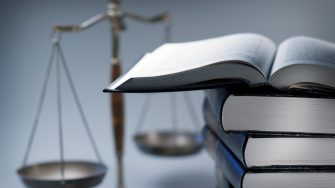 A stack of law books stands in front of a justice scale that is slightly out of focus.  On top of the stack is an open law book.