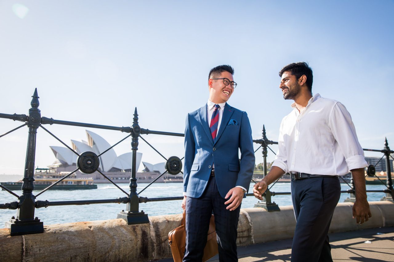 Two men walk along the Sydney Harbour foreshore in a happy discussion. The Sydney Opera House and a ferry can be seen in the background.