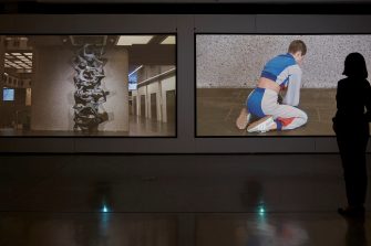 The silhouette of a person in a dark art gallery, looking at two video artworks. 