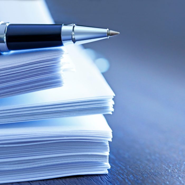 A ballpoint pen rests on top of a stack of documents ready for signing.  The image is photographed using a very shallow depth of field with the focus being on the tip of the pen.