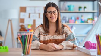 Young beautiful teacher woman wearing sweater and glasses sitting on desk at kindergarten with a happy and cool smile on face. Lucky person.