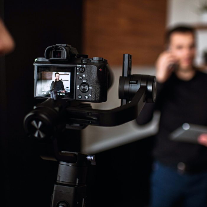 Picture with blurred background where young businessman stand in kitchen and talk on phone. Recording video of dusiness work call or meeting. Camera man stand behind camera and look to right