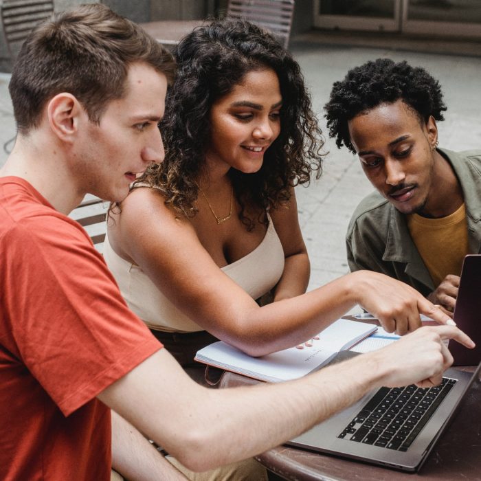 A group of three students sit together in conversation, facing an open laptop.