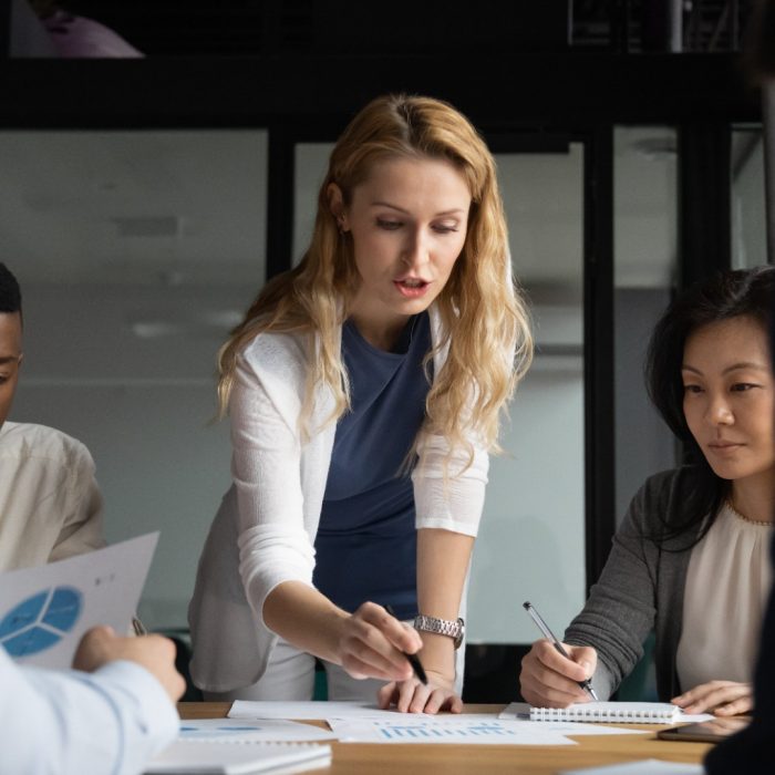 Concentrated young businesswoman explaining market research results in graphs to mixed race colleagues. Focused group of diverse employees holding brainstorming meeting, discussing project ideas.