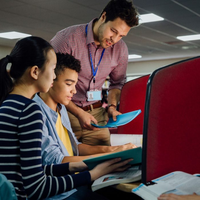 Students are working on computers with textbooks. A male teacher is standing with them, helping them with their work.