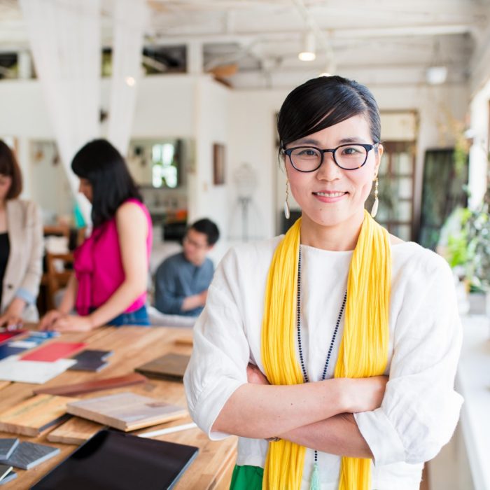 Portrait of a mid adult, female interior designer or architect looking into the camera whilst her team work away in the background.