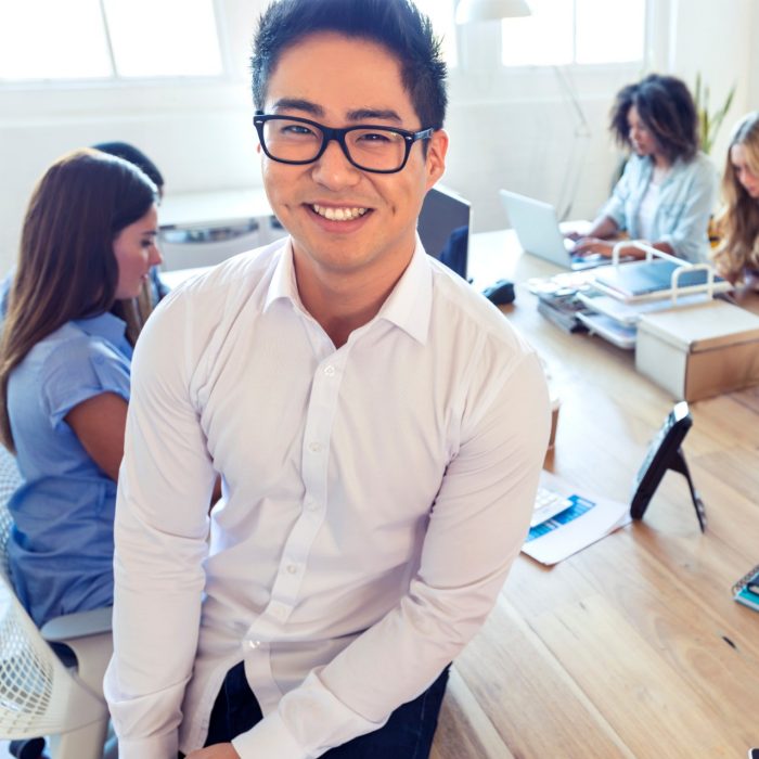 Young male professional looking at camera smiling while team work on computers in the background