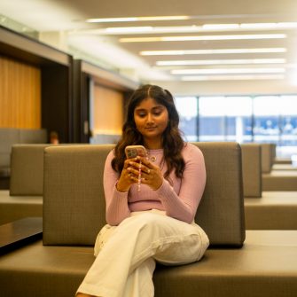 A student looking at their phone while sitting on a bench indoors on campus