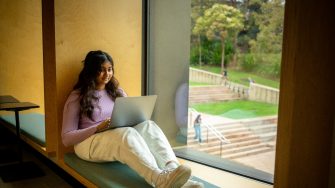 A student sitting on a window ledge at a large window with their laptop resting on their lap