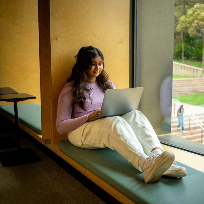 A student sitting on a window ledge at a large window with their laptop resting on their lap