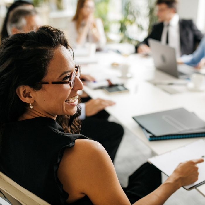 Over the shoulder profile of bespectacled female executive in early 30s sitting at conference table and laughing as she interacts with off-camera colleague.