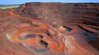 "Looking down on a large open cut iron ore mine in Australiaa's remote Pilbara region.  At the bottom of the pit at the end of the red winding road, a dump truck is being loaded with ore.  Other mine vehicles appear tiny in the huge pit.  Beyond the open cut are green hills.  Taken with permission during a guided tour of the mine."