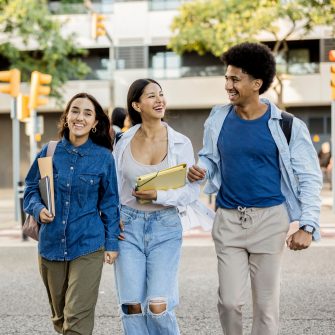 Three diverse college friends walking arm in arm, laughing and carrying books and backpacks on a sunny day.