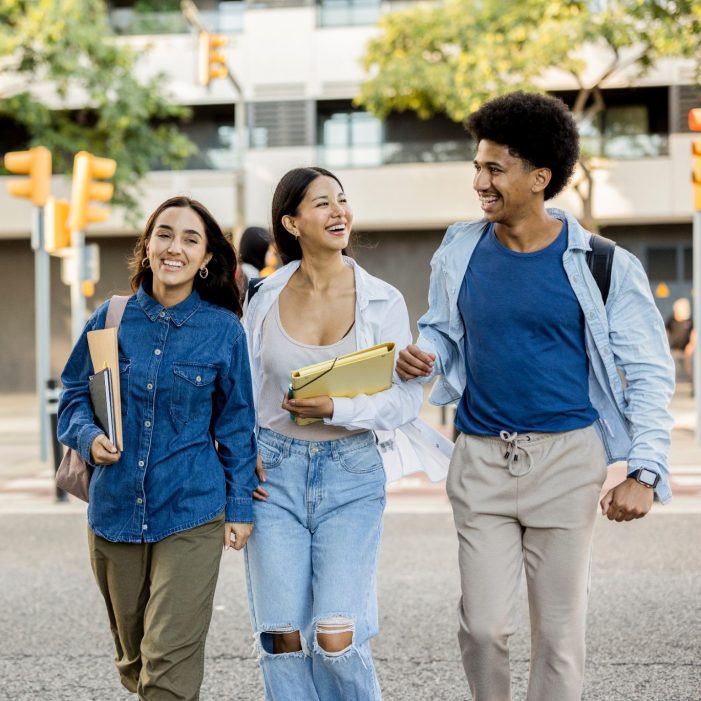 Three diverse college friends walking arm in arm, laughing and carrying books and backpacks on a sunny day.