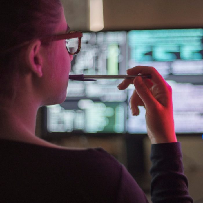 Stock image of a young woman contemplating two computer monitors, she is illuminated by the red light from a peripheral screen image.