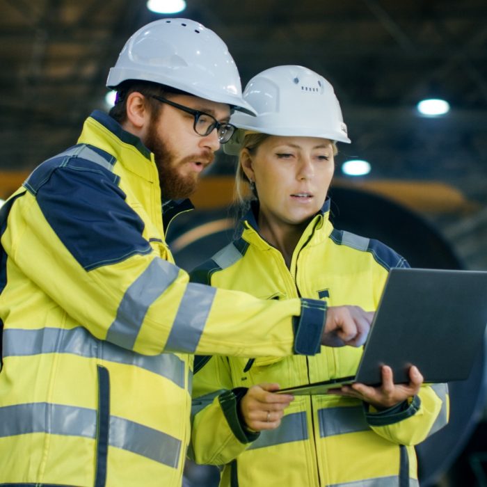 Male and Female Industrial Engineers in Hard Hats Discuss New Project while Using Laptop