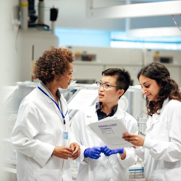 Diverse mix of technicians, wearing lab coats, debate working procedure, in a disease research facility. Nuclear magnates resonance equipment, is in the background, this high end apparatus can be used to discover information on conditions such as dementia, diabetes, autism and cancer. Focus technique and layers, ensure the group of female science graduates and manager are the centre of attention. This is a realistic scenario.