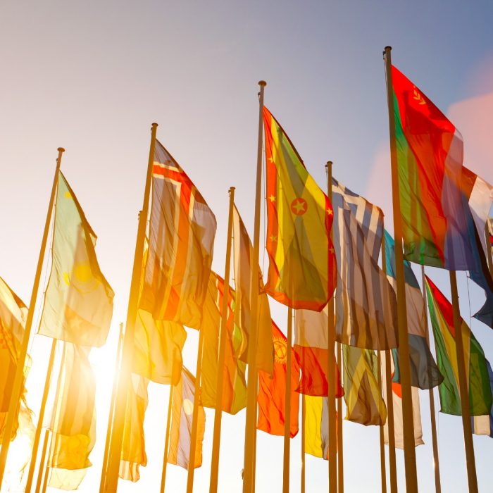 Group of many colorful internationl world flags waving in the sky at sunset, with shining sun and lens flare, low angle view.