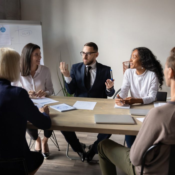 Confident male trainer holding educational workshop, sitting together with involved multiracial participants at table. Mixed race company staff listening to team leader boss ceo, explaining strategy.