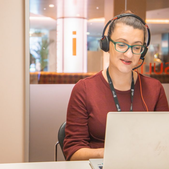 Female librarian with head set on and using laptop sits in a room with glass wall behind saying CONSULTATION in red lettering (mirror image) and orange i on white pole in distance.