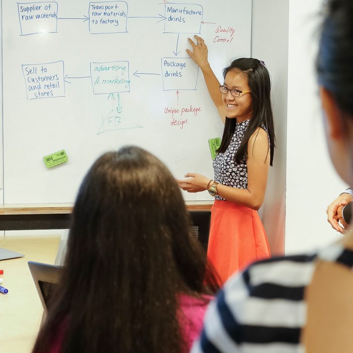 A woman standing in front of a white board pointing to diagrams on it. She is smiling at an audience who are facing towards her.