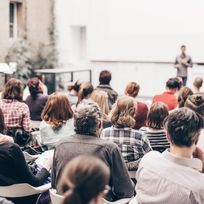 Male speaker giving presentation in lecture hall at university workshop. Audience in conference hall. Rear view of unrecognized participant in audience. Scientific conference event.