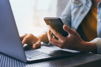 Hands typing on laptop while holding mobile phone