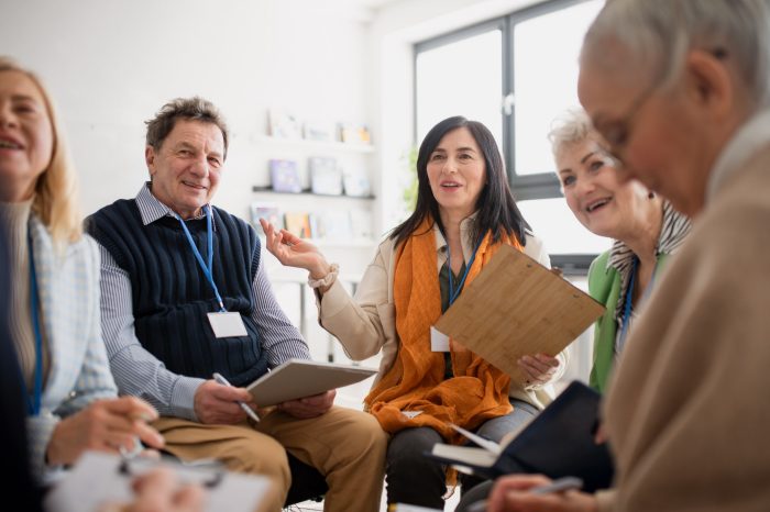 Excited elderly people attending a group therapy session at nursing house, positive senior man and woman sitting in circle, having conversation with psychologist