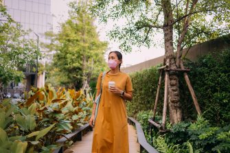 A happy Thai woman in a yellow dress carrying a cup of coffee as she walks in the park with a face mask on.