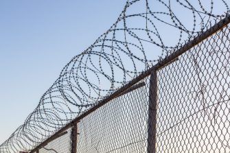 Young unidentifiable teenage boy holding hes head at the correctional institute in black and white, conceptual image of juvenile delinquency, focus on the wired fence.