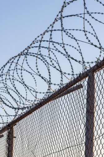 Young unidentifiable teenage boy holding hes head at the correctional institute in black and white, conceptual image of juvenile delinquency, focus on the wired fence.