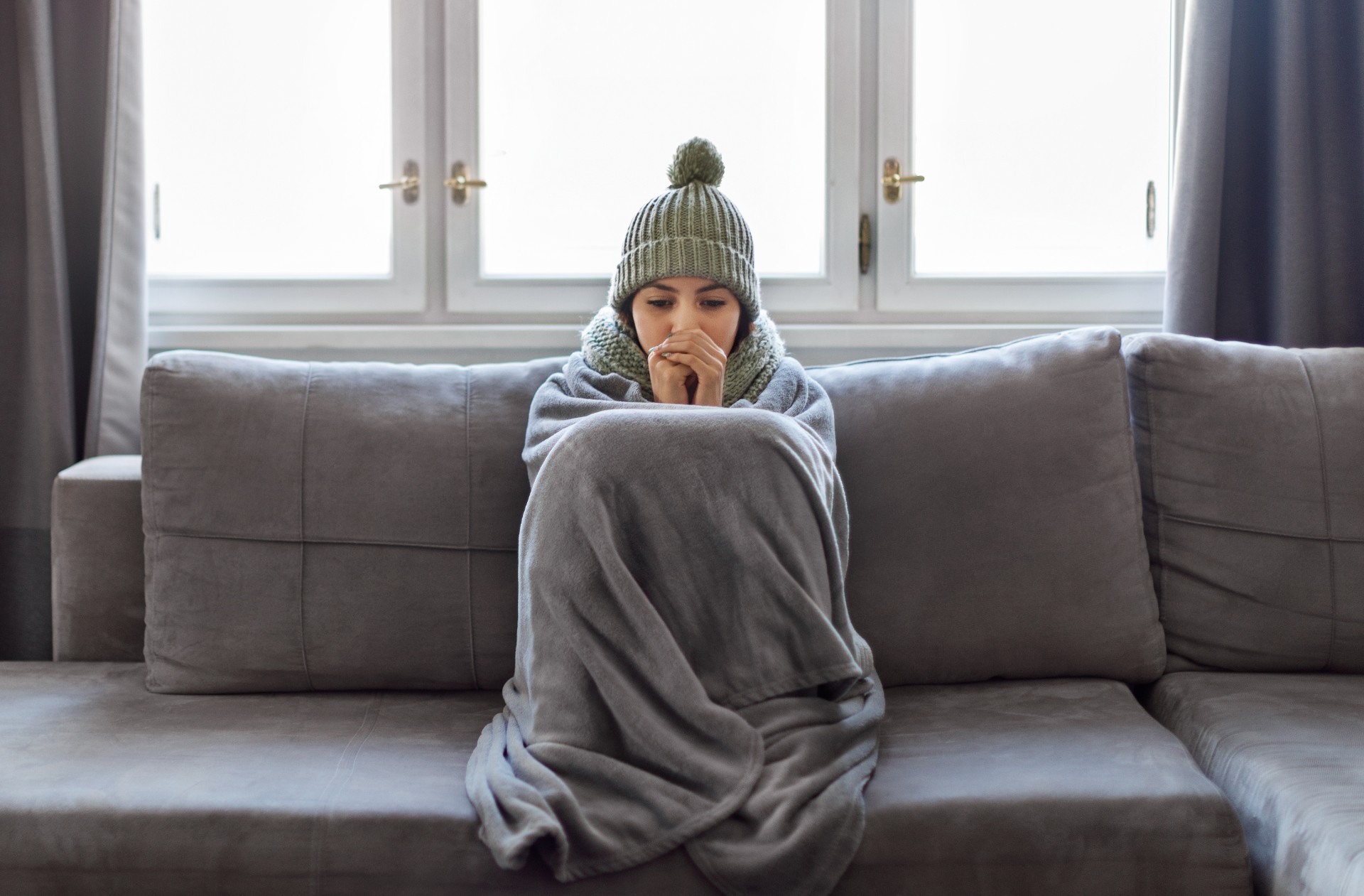 Cold Home. Young Woman Covered With Blanket Freezing On Couch In Living Room, Millennial Female Wearing Knitted Scarf And Hat Indoors Warming Hands With Her Breath, Suffering Heating Problems