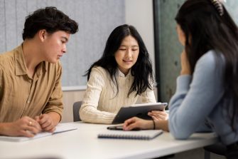 Three students sitting at a table talking over a tablet computer