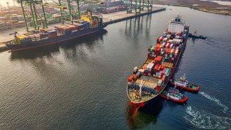 This image depicts two large shipping vessels just off port in the ocean. The image is taken from a aerial view. Each vessel has shipping containers loaded on to it.