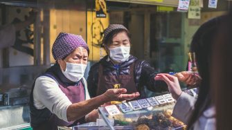 Two street vendors wearing masks are serving a customer.