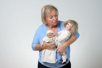 Elderly woman holding two antique dolls in her arms, conceptual of a collector or of a woman reminiscing her childhood, over grey