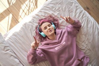 Happy funny teen girl with pink hair wear headphones lying in comfortable bed listening new pop music enjoying singing song with eyes closed relaxing in cozy bedroom at home. Top view from above.