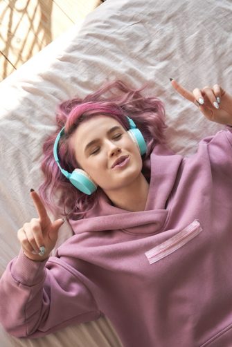 Happy funny teen girl with pink hair wear headphones lying in comfortable bed listening new pop music enjoying singing song with eyes closed relaxing in cozy bedroom at home. Top view from above.