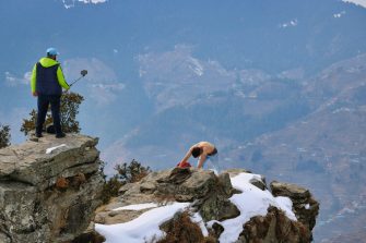 A man poses on the edge of a cliff as another man takes his photo from a rocky outcrop above.