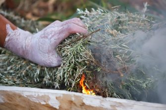 A hand covered in white ceremonial mud places branches into flame to create fire