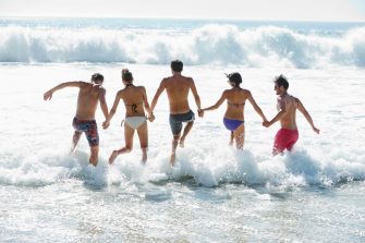 Four young people in swimmers walk through foamy waves on the beach towards the biggers waves deeper out.