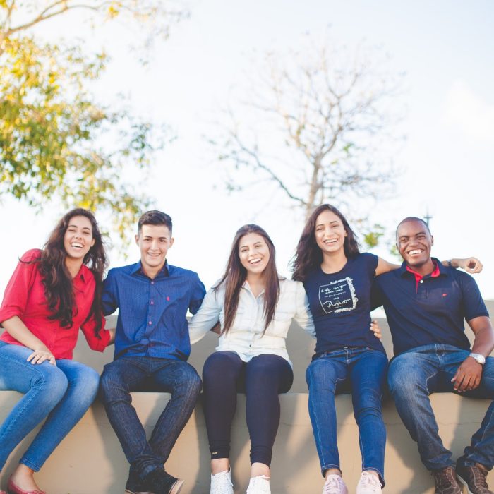 Five happy people sitting on a bench with trees and a blue sky