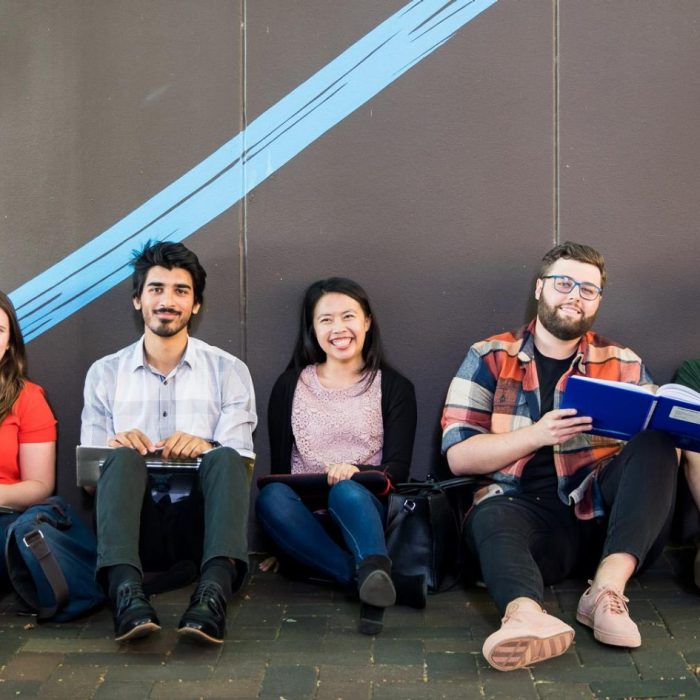 Five students sitting on the ground against a wall smiling