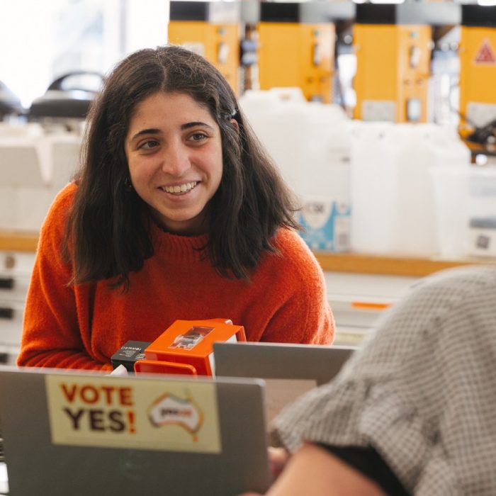 Dark haired woman in orange jumper smiling at another person who is facing her