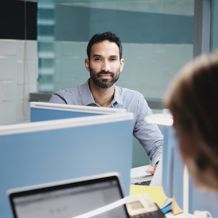 Portrait of serious male manager working in modern office with female colleagues. Busy hispanic business man leaning on desk at work in executive coworking space, looking at camera.