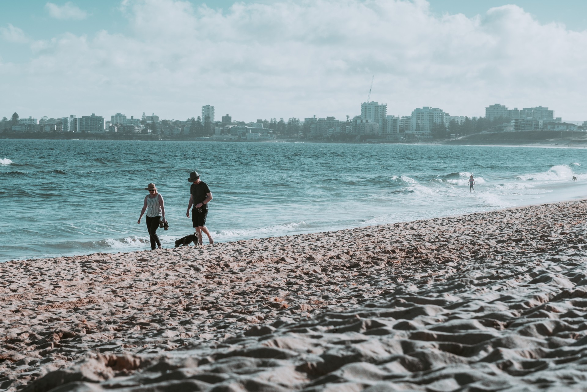 Two people stroll along a beach with city skyline in the background