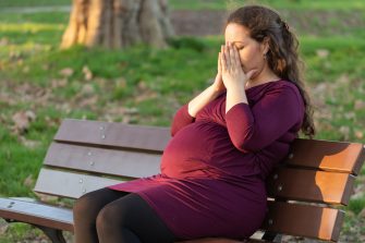 Young pregnant woman taking a moment to de-stress and relax as she suffers morning sickness seated in warm sunshine on a park bench with her hands covering her face and eyes closed