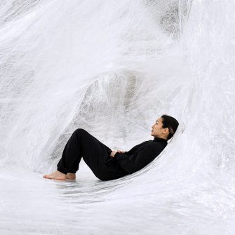 A woman in black clothing reclines within a giant web of woven white fibres like cotton wool