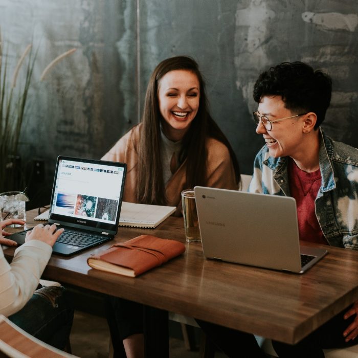 Three women laughing while sitting at a table with laptops