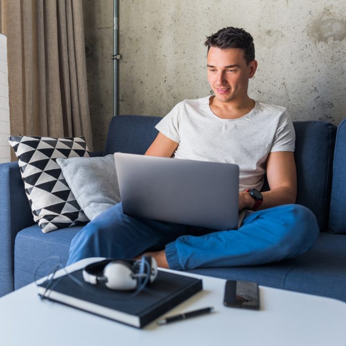 Young man sitting on sofa at home working on laptop online, using internet, smiling